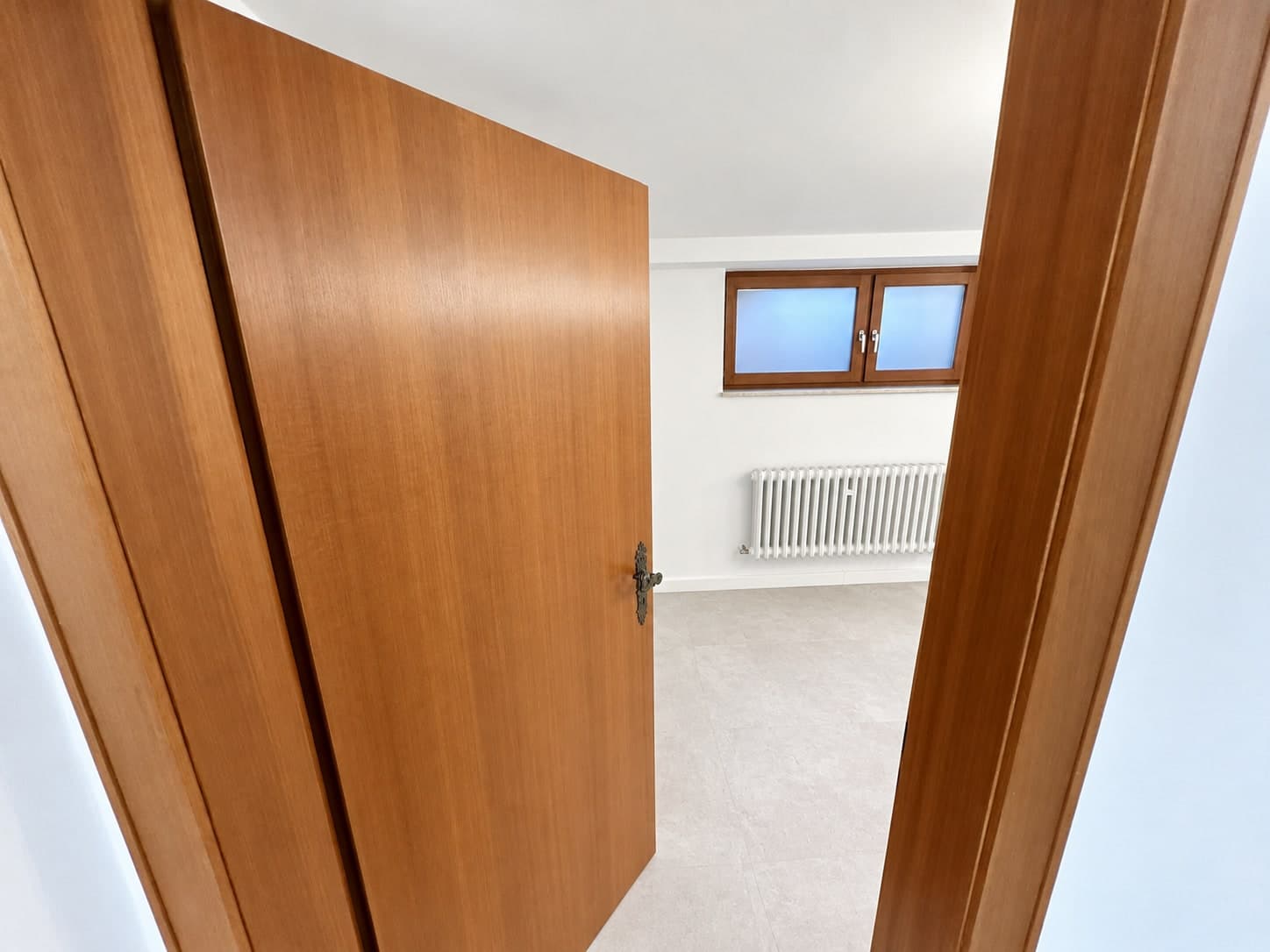 Blick durch eine teilweise geöffnete Holztür auf ein hohes Fenster und einen Heizkörper