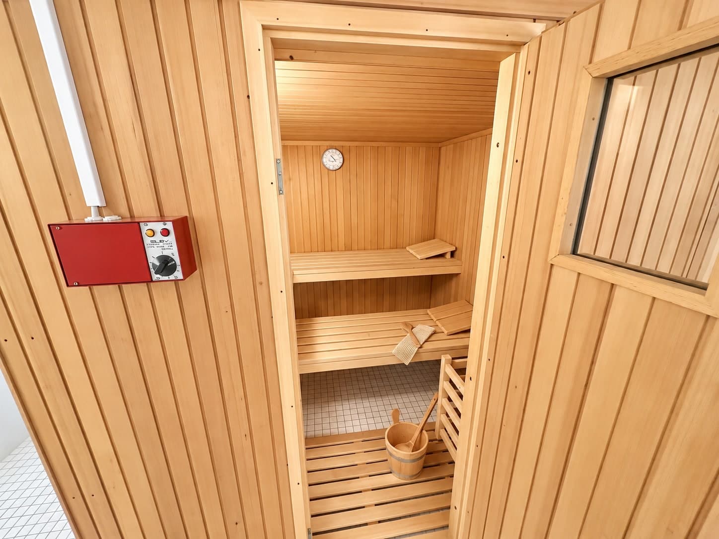 Holzsauna mit Sitzbänken, Saunakübel und roter Steuerung neben der Tür